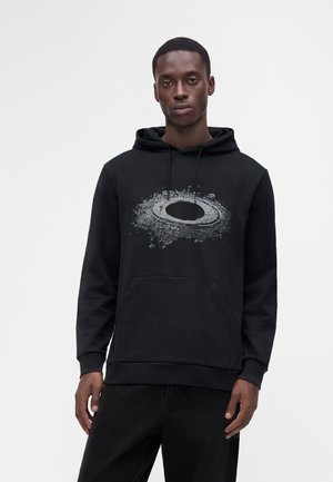Jeune homme portant un sweat à capuche noir avec un motif gris sur le thème de l'espace représentant une planète avec des anneaux sur le devant, debout devant un fond uni.
