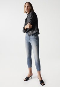 Schwarze Lederjacke, hellblaue, ausgebleichte Jeans, offene schwarze High Heels. Slim Fit, cropped Länge, leichte Ausbleichungen an den Jeans, minimale Verzierungen.