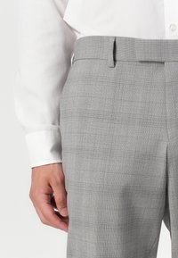 Pantaloni a quadri grigi realizzati in un tessuto testurizzato. Presentano una parte superiore piatta, passanti per la cintura e una vestibilità su misura. Indossati con una camicia bianca a maniche lunghe.
