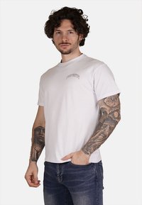 T-shirt bianco in cotone con colletto rotondo e maniche corte, dotato di un piccolo logo grafico testuale sulla parte alta sinistra del petto.
