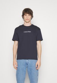 Navy zila kokvilnas T-kreklu ar apaļu kaklu, kas rotāts ar baltu "Calvin Klein" logo uz krūtīm. Īsas piedurknes un brīva piegriezuma dizains.