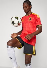 Mannelijke voetballer in rood-zwart tenue die een witte bal met zwarte sterren op zijn knie balanceert tegen een effen achtergrond.