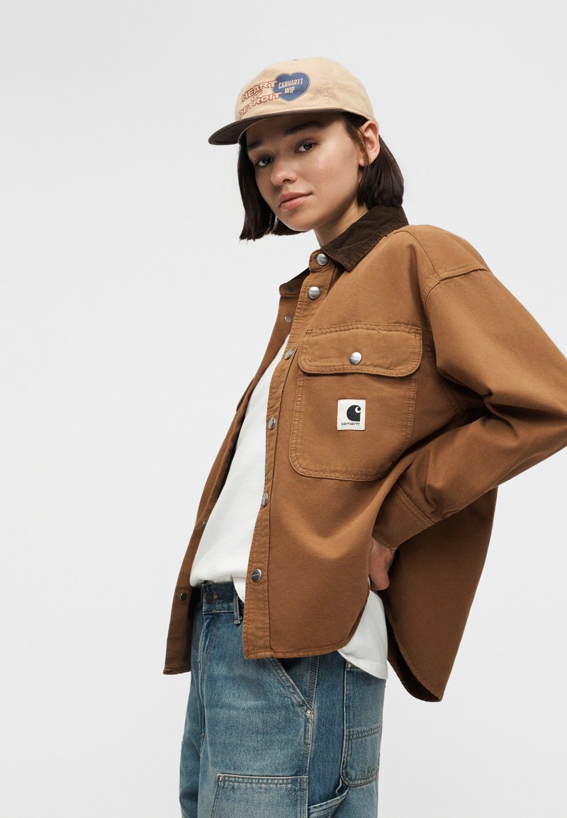 Giovane che indossa una giacca Carhartt color cuoio, maglietta bianca, jeans blu e un berretto beige con toppe, posando con la mano sui fianchi davanti a uno sfondo semplice.