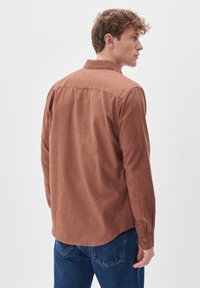 Camicia marrone a maniche lunghe realizzata in tessuto texturizzato; presenta un colletto e polsini con bottoni, un orlo curvo e un dettaglio di yoke sul retro.