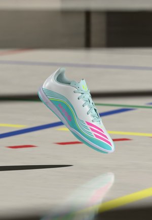 White and teal indoor soccer shoe with pink "MESSI" text and logo, floating above a multi-colored indoor sports court floor.