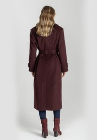 Długi burgundowy płaszcz z paskiem w talii, dopasowanym kroju i bocznymi rozcięciami. Noszony na dopasowane dżinsy i kozaki za kostkę. Gładka tekstura materiału.
