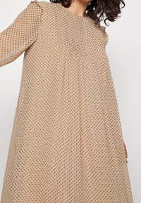 Langärmliges, beiges Kleid mit braunen Punkten. Es hat ein gerafftes Joch und Rüschendetails an den Schultern und besteht aus leichtem, strukturiertem Stoff.