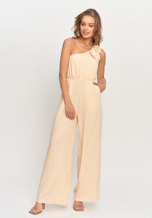 Kobieta ubrana w beżowy kombinezon bez rękawów z szerokimi nogawkami i kokardą na jednym ramieniu, stojąca z jedną ręką w kieszeni.