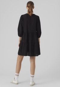 Vestido negro con volantes y mangas de tres cuartos abullonadas, hecho de una tela ligera. Combinado con zapatillas blancas y calcetines a rayas.