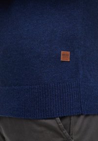 Navy blue neulepaita, jossa on ribbettä alareunassa ja pieni ruskea nahkalaatta, jossa on teksti "INDICODE EST.1993" edessä.