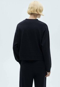 Personne aux cheveux blonds courts portant un haut noir ample à manches longues et un pantalon assorti, vue de dos sur un fond uni.