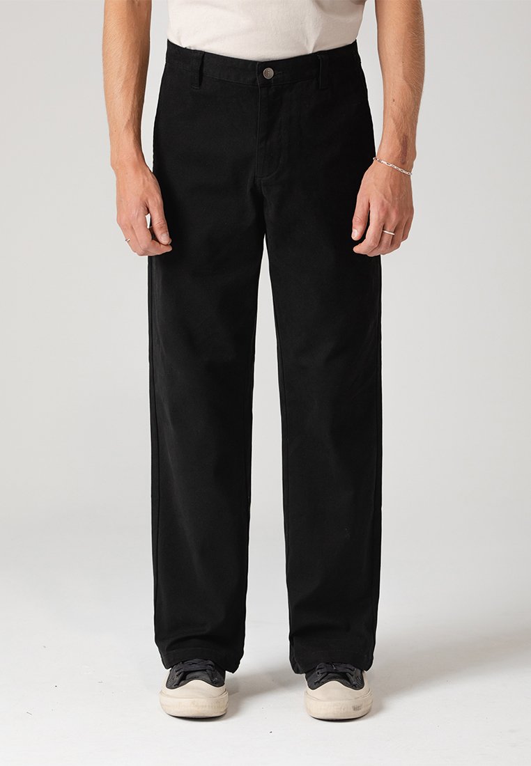 Μαύρο βαμβακερό παντελόνι με ίσια γραμμή, διαθέτει μπροστινό κούμπωμα με κουμπί και �πλαινές τσέπες, φορεμένο με μαύρα αθλητικά παπούτσια.