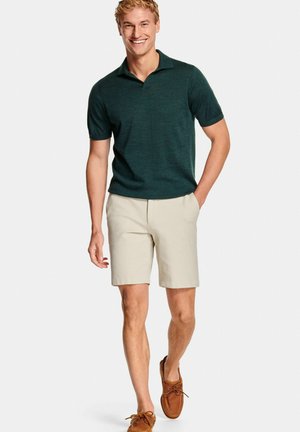 Man walking wearing dark green short-sleeve polo, beige shorts, and brown moccasin shoes, smiling with one hand in pocket.