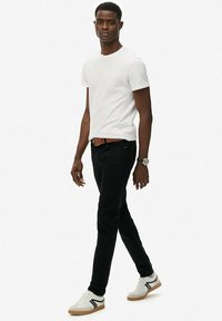 Camiseta blanca de algodón, jeans negros, cinturón marrón y zapatillas blancas con detalles en gris y negro. El modelo está de pie en un fondo neutro.