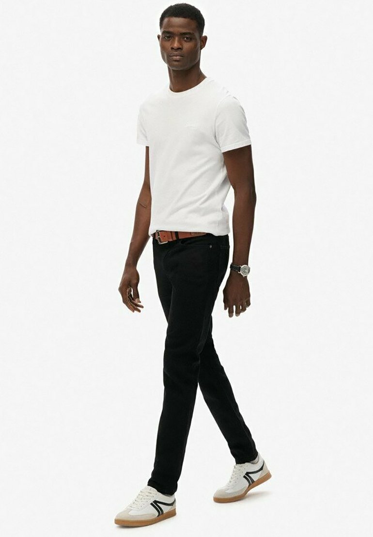 Camiseta blanca de algodón, jeans negros, cinturón marrón y zapatillas blancas con detalles en gris y negro. El modelo está de pie en un fondo neutro.