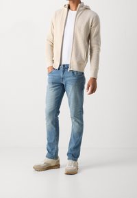 Beige zip-up hoodie with textured fabric, white t-shirt, light blue jeans, and beige sneakers with grey accents. Standing posture, hands in pockets.