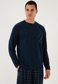 Tamnoplava košulja dugih rukava s karirastim džepom, okrugli izrez i mekanim teksturom, uparen s karirastim hlačama koje imaju svijetloplave i bijele pruge.