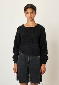 Juodas pūkuotas megztinis su apykakle ir ilgomis, šiek tiek puffuotomis rankovėmis, derinamas su laisvomis juodomis džinsinėmis šortais su nelygiais kraštais.