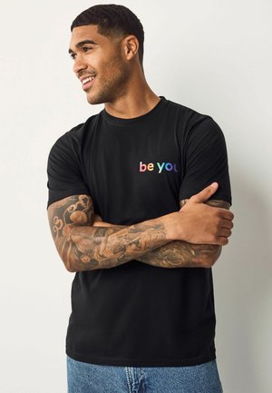 Sort t-shirt med "be you" tekst i regnbuens farver på venstre side. Korte ærmer, rund hals, lavet af blødt stof. Casual pasform.