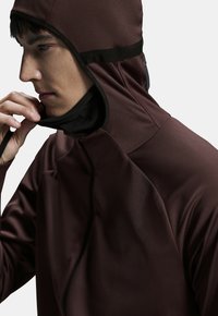 Giacca marrone aderente con collo alto e cappuccio, caratterizzata da un tessuto testurizzato e accenti neri. Dettaglio con zip visibile sul davanti.