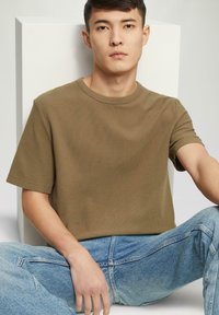 Oliwkowa koszulka z krótkim rękawem o teksturowanej tkaninie, okrągłym dekolcie i luźnym kroju, w połączeniu z jasnoniebieskimi dżinsami.