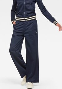 Marinblå atletisk träningsdräkt med vida byxor och en zip-up jacka med horisontella randade detaljer på ärmarna. Mjuk, andningsbar tyg med logga.