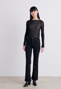 Haut noir en tissu transparent à manches longues, ajusté à la taille, associé à un jean noir évasé taille haute. Le mannequin porte des boots noires à talons aiguille.