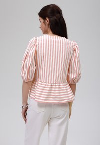 Pfirsichfarbener und weiß gestreifter Bluse mit Puffärmlen, Peplum-Saum und einem Schlüsselhetz-Design an der Rückseite, kombiniert mit weißen Jeans. Glatte Stoffoberfläche.