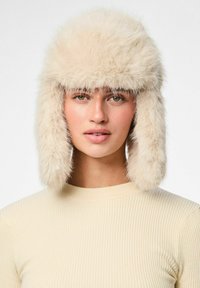 Chapeau en fausse fourrure de couleur blanc cassé avec un dessus duveteux et des rabats pour les oreilles. Texture douce, design arrondi, complète un pull à côtes légères.