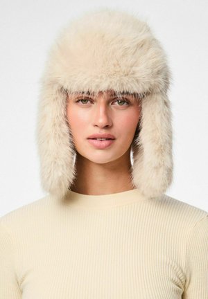 Faux-fur muts in off-white met een fluffy bovenkant en oorkleppen. Zachte textuur, afgeronde vorm, het complimenteert een licht geribbeld sweater.