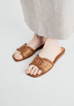 Fødder iført brune, åbne, flettede slip-on sandaler med spænde-detalje, kombineret med beige linnedbukser.