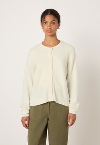 Young woman wearing a cream buttoned cardigan and olive green pants, standing against a plain light background.