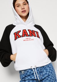 Hanorac cu glugă alb și negru, cu broderie roșie "KANI", având un buzunar frontal și șnur, asortat cu pantaloni albaștri cu model.