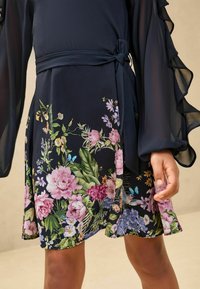Rochie cu imprimeu floral, cu un top din voal de un albastru marin închis, cu mâneci volan. Prezintă un cordon din același material. Are motive florale vibrante în nuanțe de roz, violet și verde la tiv.