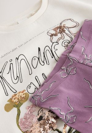 Lysecreme sweatshirt med blomster-sequin-designs og sort tekst, kombineret med lilla bukser med et lignende blomstermønster og tekstur.