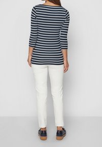 Mujer vistiendo una blusa de rayas horizontales en azul marino y blanco de manga tres cuartos, pantalones blancos con patrones sutiles y zapatos de plataforma azul marino, de pie.