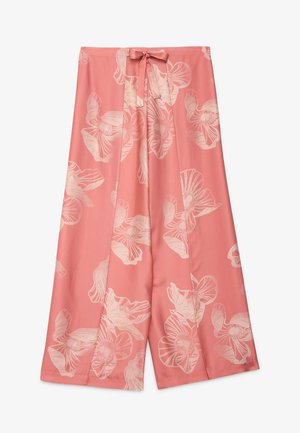 Femme aux cheveux lissés portant un haut rose ample noué à la taille et une jupe longue rose à fleurs, debout devant un fond clair.