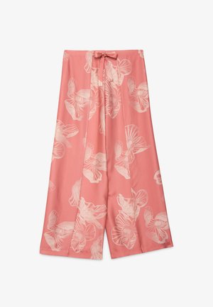 Kvinde med glat frisure bærer en løs pink top bundet i taljen og en flydende pink blomsterskørt, stående mod en lys baggrund.