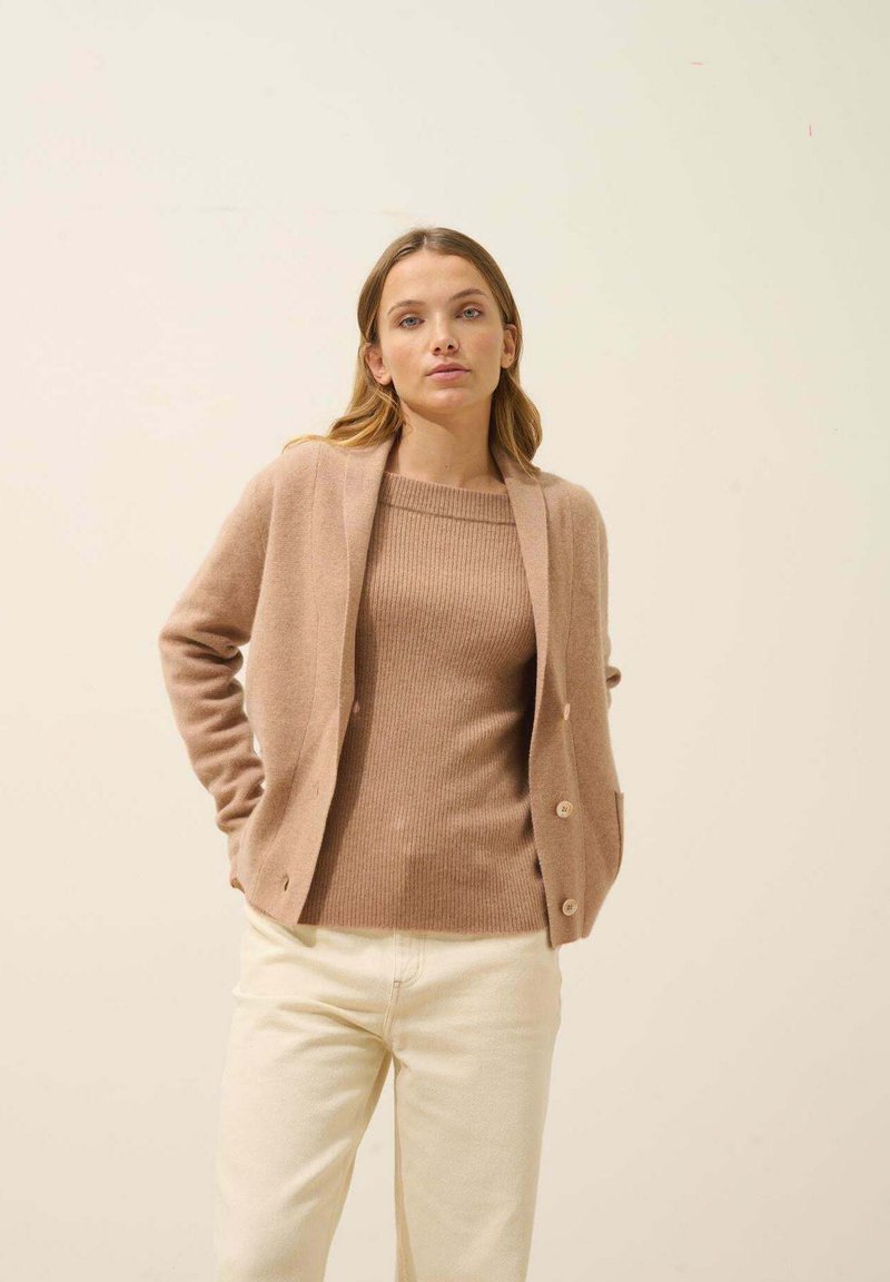 Cárdigan beige con cierre de botones y mangas largas sobre un suéter marrón con textura en relieve, combinado con pantalones color crema. Textura suave en toda la prenda.