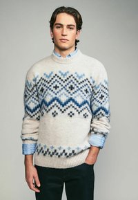 Pulover din lână bej deschis, cu modele geometrice albastre și negre. Terminatii și mansete ribbate. Poartă-l peste o cămașă cu dungi albastre deschise.
