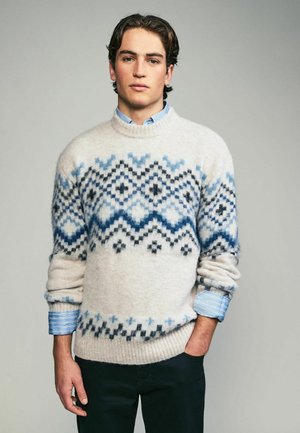 Light beige wool sweater with blue and black geometric patterns. Ribbed hem and cuffs. Layered over a light blue striped shirt.