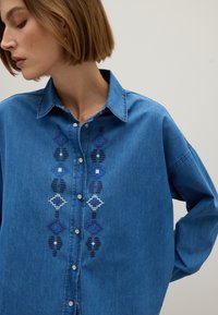 Frau, die ein blaues Denim-Hemd mit vertikaler geometrischer Stickerei in verschiedenen Blautönen und Weiß trägt und nach unten zur Seite schaut.