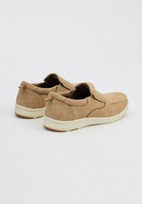 Zapatos de ante color tostado, tipo slip-on, con un acabado texturizado, suela de goma y lengüeta en la parte trasera. El diseño presenta costuras mínimas y una silueta de perfil bajo.
