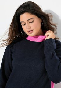 Pull en maille bleu marine avec un col rose à côtes. Le tissu semble doux, avec des manches longues et des détails texturés subtils.