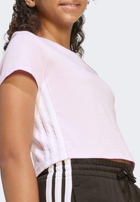 Svijetlo ružičasta skraćena majica s kratkim rukavima i bijelim bočnim trakama, u kombinaciji s crnim hlačama koje imaju tri bijele trake. Glatka tekstura tkanine.