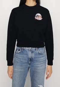 Sudadera negra recortada con puños y cuello acanalados. Presenta un parche gráfico colorido de dos personajes en un marco circular en el pecho.