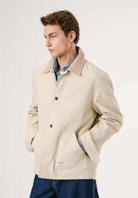 Joven con chaqueta beige con cuello sobre camisa azul y jeans, manos en los bolsillos, mirando de lado contra un fondo liso.