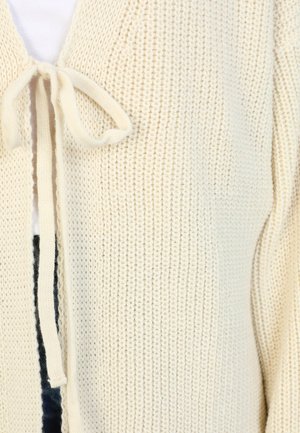 Cream-colored knitted cardigan with a textured finish, featuring a tie closure at the neckline and open front design.
