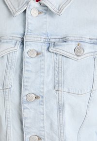 Světle modrá denimová bunda s kovovými knoflíky, dvěma náprsními kapsami a kontrastním prošíváním. Hladká textura s vybledlými místy.