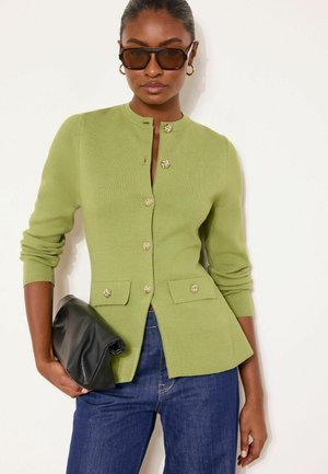 Žena v olivovo zelenej gombíkovej karidgane, modrých džínsoch, tmavých slnečných okuliaroch, zlatých kruhových náušniciach, držiaca čiernu listovú kabelku.